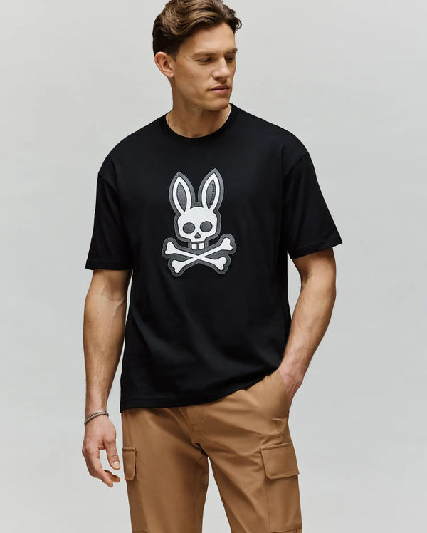 A man wearing the Psycho Bunny MENS RYDER OVERSIZED FIT TEE (MT0200062), featuring an embroidered Bunny skull and crossbones graphic, stands with one hand in his tan cargo pants pocket, gazing slightly to the side against a plain backdrop.