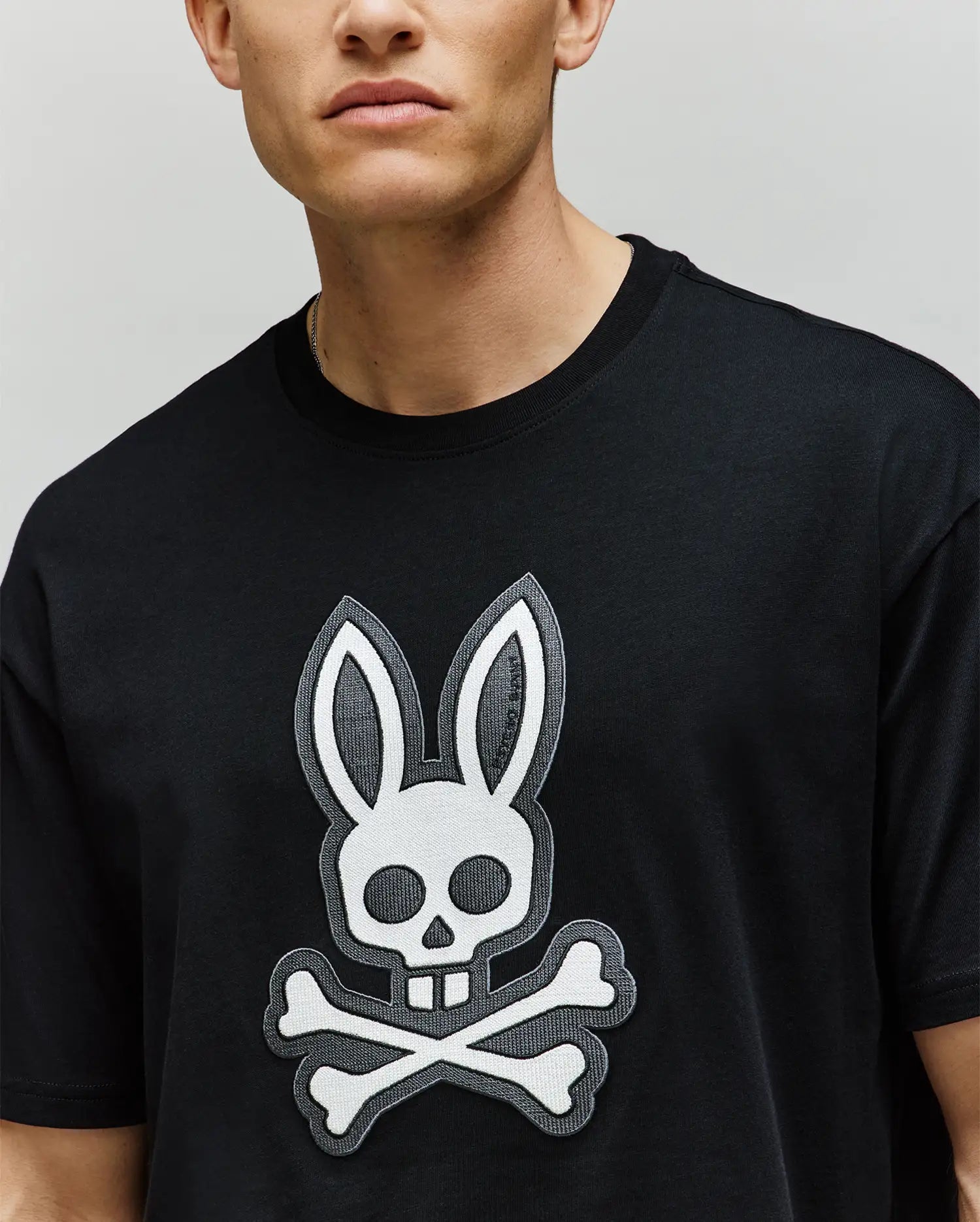 A person wears the Psycho Bunny MENS RYDER OVERSIZED FIT TEE (MT0200062), a black premium shirt featuring a bold white bunny skull and crossbones graphic on the front; only the lower half of their face is shown.