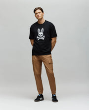 A man with short hair and a relaxed posture wears the Psycho Bunny MENS RYDER OVERSIZED FIT TEE (MT0200062), featuring premium Pima cotton and an embroidered Bunny skull, paired with tan cargo pants and black Adidas sneakers.