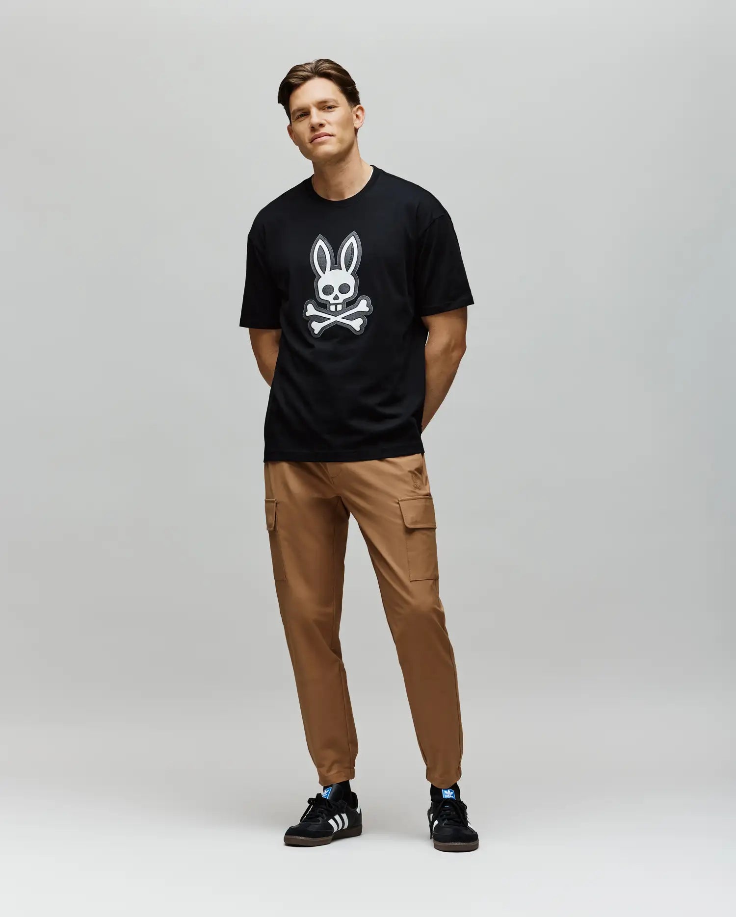 A man with short hair and a relaxed posture wears the Psycho Bunny MENS RYDER OVERSIZED FIT TEE (MT0200062), featuring premium Pima cotton and an embroidered Bunny skull, paired with tan cargo pants and black Adidas sneakers.