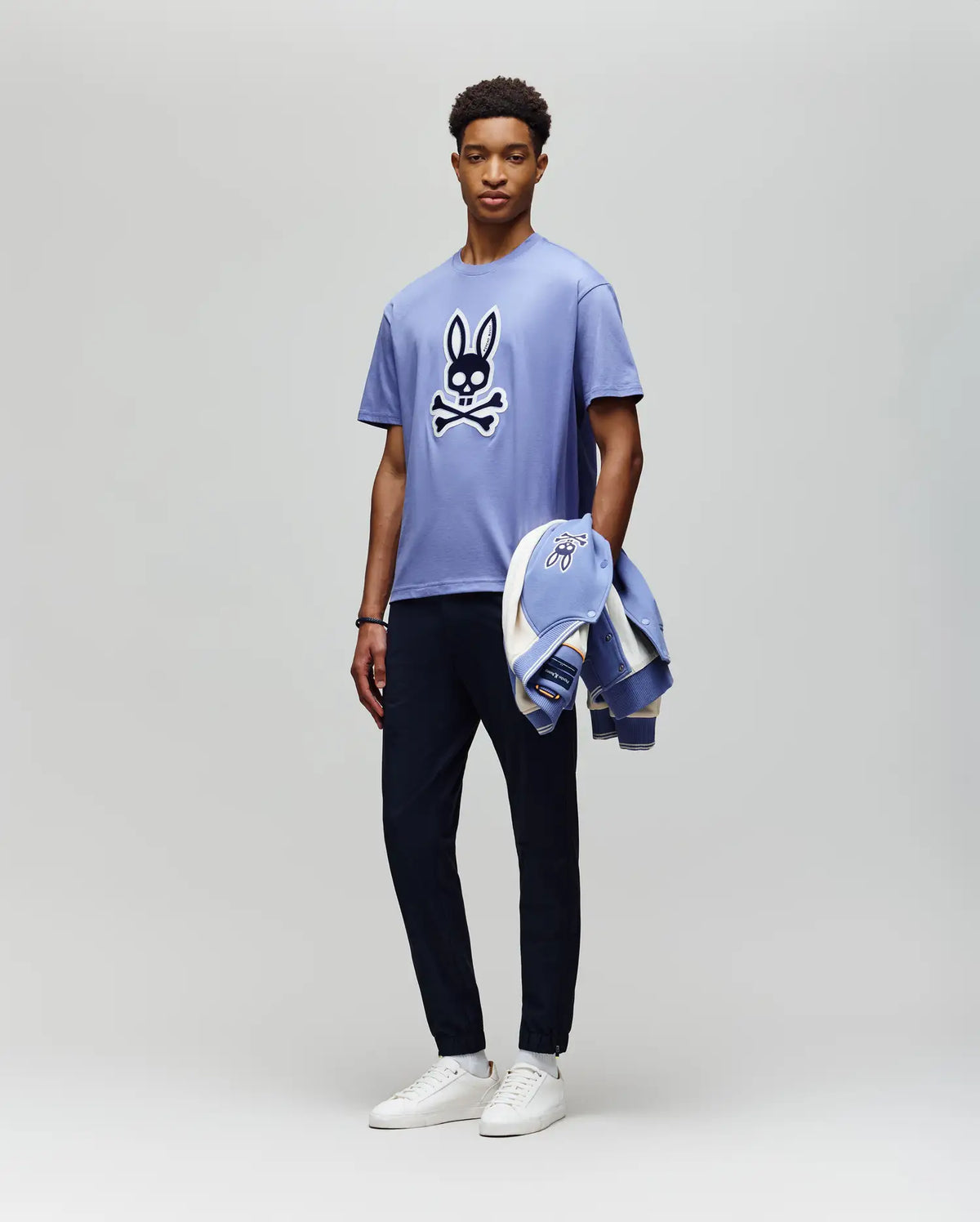 A man stands against a plain background wearing the Psycho Bunny MENS RYDER OVERSIZED FIT TEE (MT0200062), a light purple premium Pima cotton shirt with an embroidered Bunny skull and crossbones, dark pants, white sneakers, and holds a matching jacket.