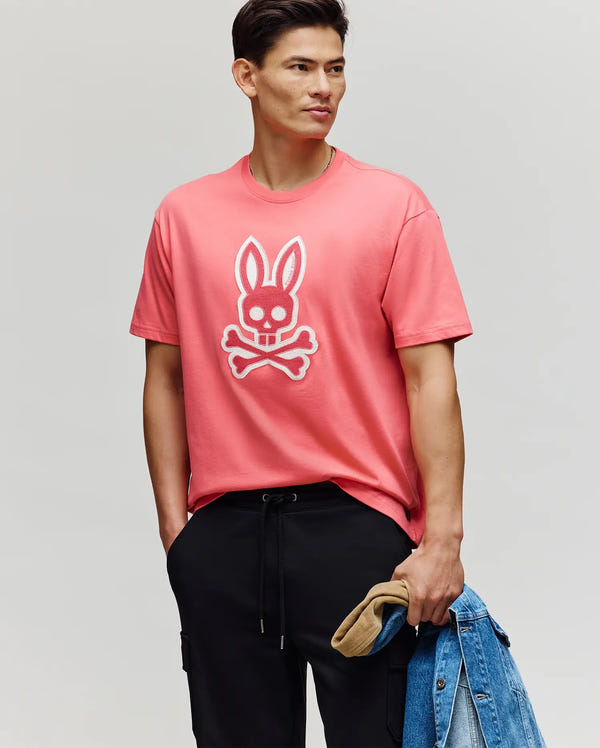 A man wears the Psycho Bunny MENS RYDER OVERSIZED FIT TEE (MT0200062) in pink, featuring an embroidered Bunny skull and crossbones. He pairs it with black pants, holds a denim jacket and tan cap, and stands with a hand in his pocket.