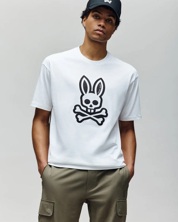 A man in a black cap, olive green cargo pants, and a Psycho Bunny MENS RYDER OVERSIZED FIT TEE—featuring an embroidered Bunny skull and crossbones—stands with hands in pockets against a plain light gray background.