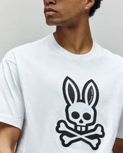 A person looks to the side, shown from the shoulders up, wearing a Psycho Bunny MENS RYDER OVERSIZED FIT TEE (MT0200062), a white Pima cotton shirt with a large black skull-and-crossbones emblem featuring rabbit ears on the front.