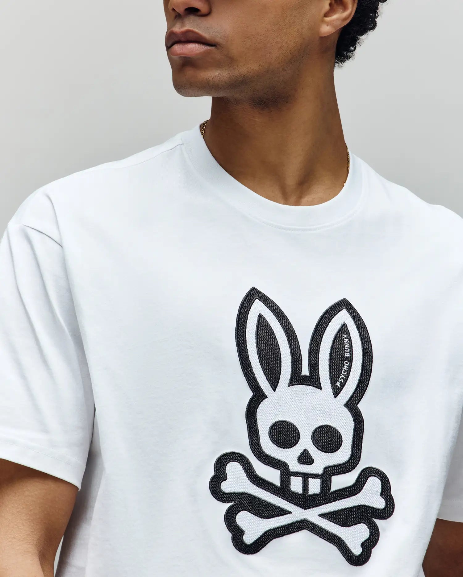 A person looks to the side, shown from the shoulders up, wearing a Psycho Bunny MENS RYDER OVERSIZED FIT TEE (MT0200062), a white Pima cotton shirt with a large black skull-and-crossbones emblem featuring rabbit ears on the front.