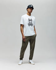 A man stands against a plain background wearing the Psycho Bunny MENS RYDER OVERSIZED FIT TEE (MT0200062) with a large black skull and embroidered Bunny graphic, olive green pants, white sneakers, and a black cap. His hands are in his pockets.