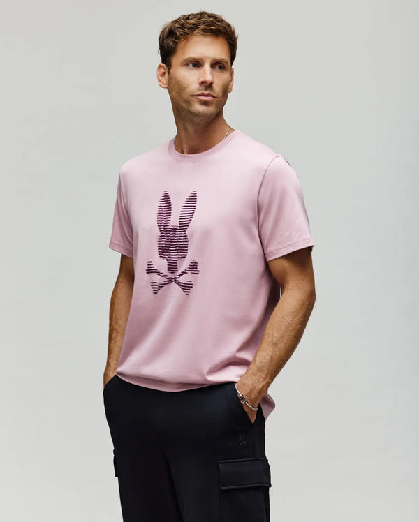A man wears the Psycho Bunny MENS GRANT BUNNY GRAPHIC TEE (MT0200081), a light pink Pima cotton shirt with a stylized bunny and crossbones, paired with black pants, standing hands-in-pockets against a plain light background.