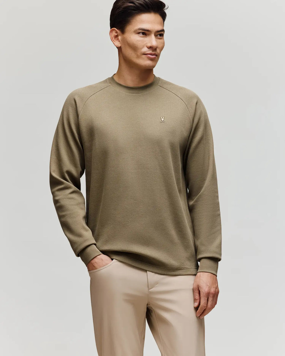 A man with short, styled hair and a slight smile stands against a plain background, wearing Psycho Bunny’s MENS EVANS WAFFLE - MT0200091 khaki cotton knit crewneck sweater and light beige pants, one hand in his pocket.