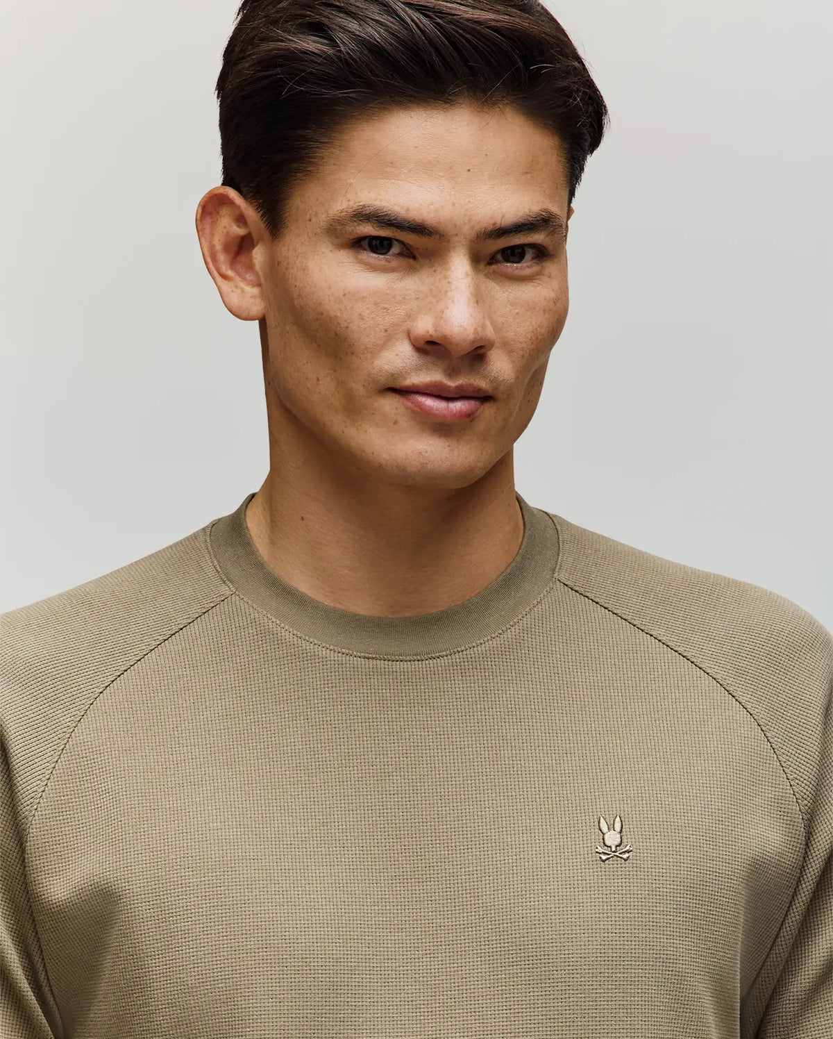 A man with short dark hair wears the Psycho Bunny MENS EVANS WAFFLE - MT0200091, a light olive green cotton crew-neck sweater with an embroidered Bunny logo on the left chest, standing against a plain light background.