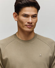 A man with short dark hair wears the Psycho Bunny MENS EVANS WAFFLE - MT0200091, a light olive green cotton crew-neck sweater with an embroidered Bunny logo on the left chest, standing against a plain light background.
