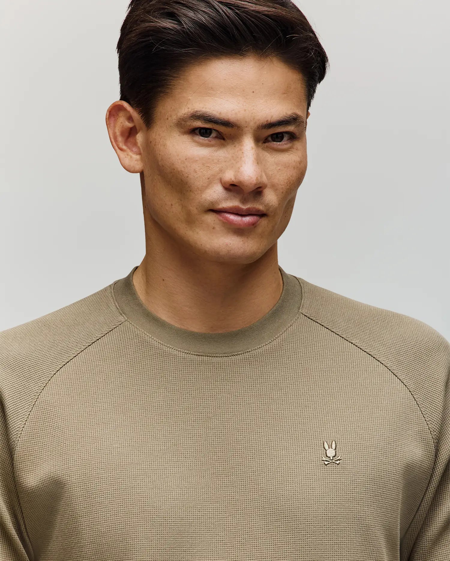A man with short dark hair wears the Psycho Bunny MENS EVANS WAFFLE - MT0200091, a light olive green cotton crew-neck sweater with an embroidered Bunny logo on the left chest, standing against a plain light background.