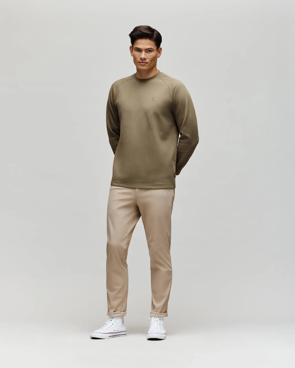A man poses against a plain background in the Psycho Bunny MENS EVANS WAFFLE - MT0200091 olive green cotton knit long-sleeve shirt, beige pants, and white sneakers, with his hands behind his back and looking slightly to the side.