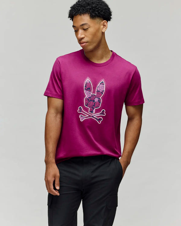 A man stands against a plain gray background wearing the Psycho Bunny MENS ROWEN BUNNY GRAPHIC TEE (MT0200101), a magenta Pima cotton shirt with a bunny skull and crossbones graphic, one hand in his pocket as he looks down to the side.