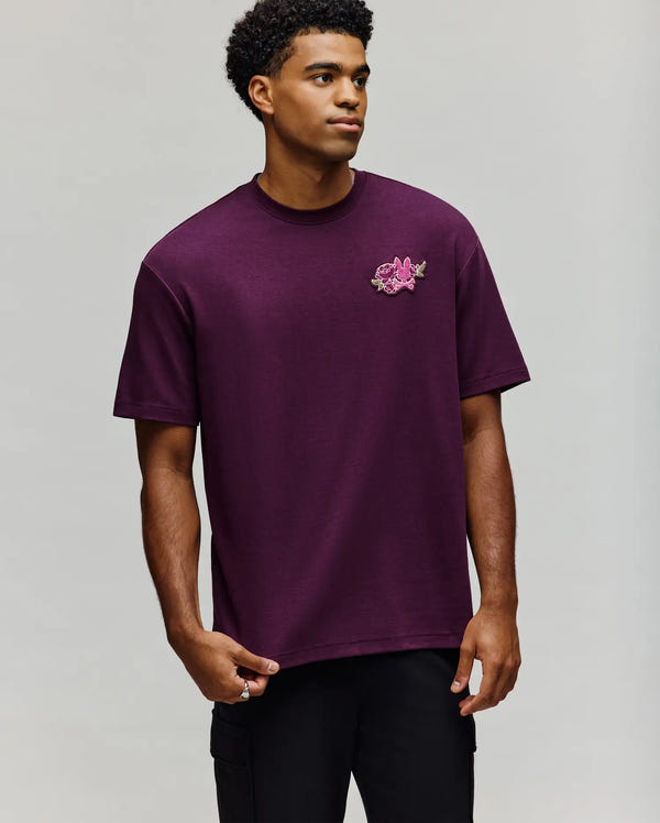 A young man models the Psycho Bunny MENS AUGUSTE OVERSIZED TEE (MT0200104) in dark purple, featuring a relaxed fit and pink floral chest embroidery, standing against a light grey background with effortless style.