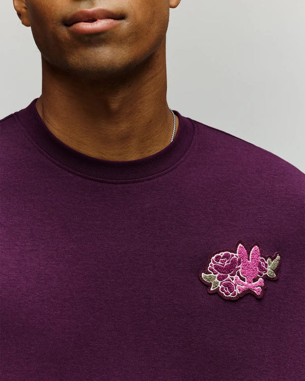 A man wears the Psycho Bunny MENS AUGUSTE OVERSIZED TEE (MT0200104), a purple crewneck tee featuring a small pink flower and bird embroidery on the left chest. Only his lower face and upper torso are visible, giving off a relaxed vibe.
