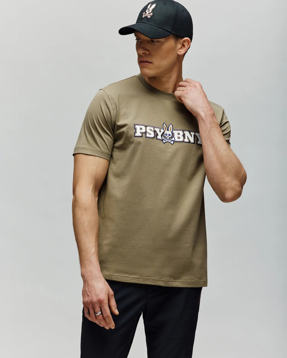 A man in a black cap and the Psycho Bunny MENS WADE GRAPHIC TEE (MT0200105)—olive-green with "PSYBUNY" and a cartoon bunny graphic—stands against a light gray background, looking sideways with one hand on his neck.