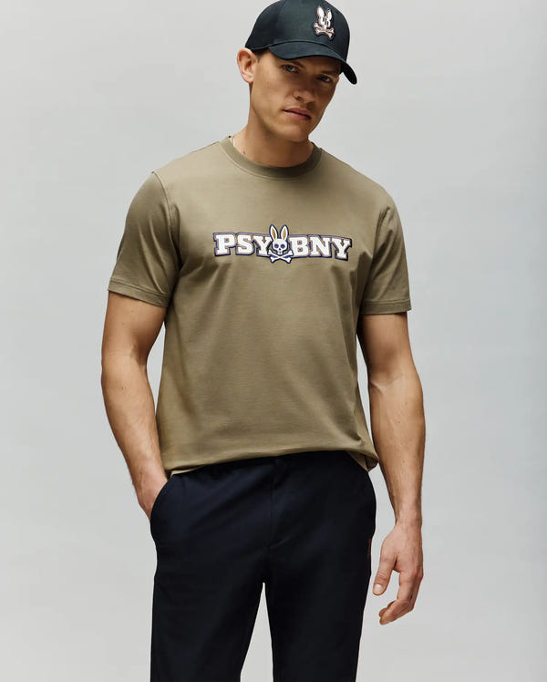 A man in a regular fit, 100% cotton olive green MENS WADE GRAPHIC TEE - MT0200105 by Psycho Bunny with a "PSYBNY" and cartoon bunny logo stands with one hand in his pocket, wearing a black cap against a light gray background.