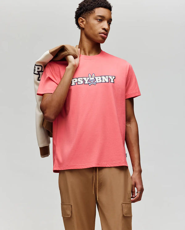 A young man wears the Psycho Bunny MENS WADE GRAPHIC TEE - MT0200105, a regular fit 100% cotton pink tee featuring "PSYBNY" and a cartoon bunny, paired with tan cargo pants and holding a beige and brown jacket.