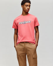 A person wears the Psycho Bunny MENS WADE GRAPHIC TEE - MT0200105, a coral pink 100% cotton tee with "PSYBNY" and a cartoon bunny on the chest, paired with tan cargo pants against a light gray background.