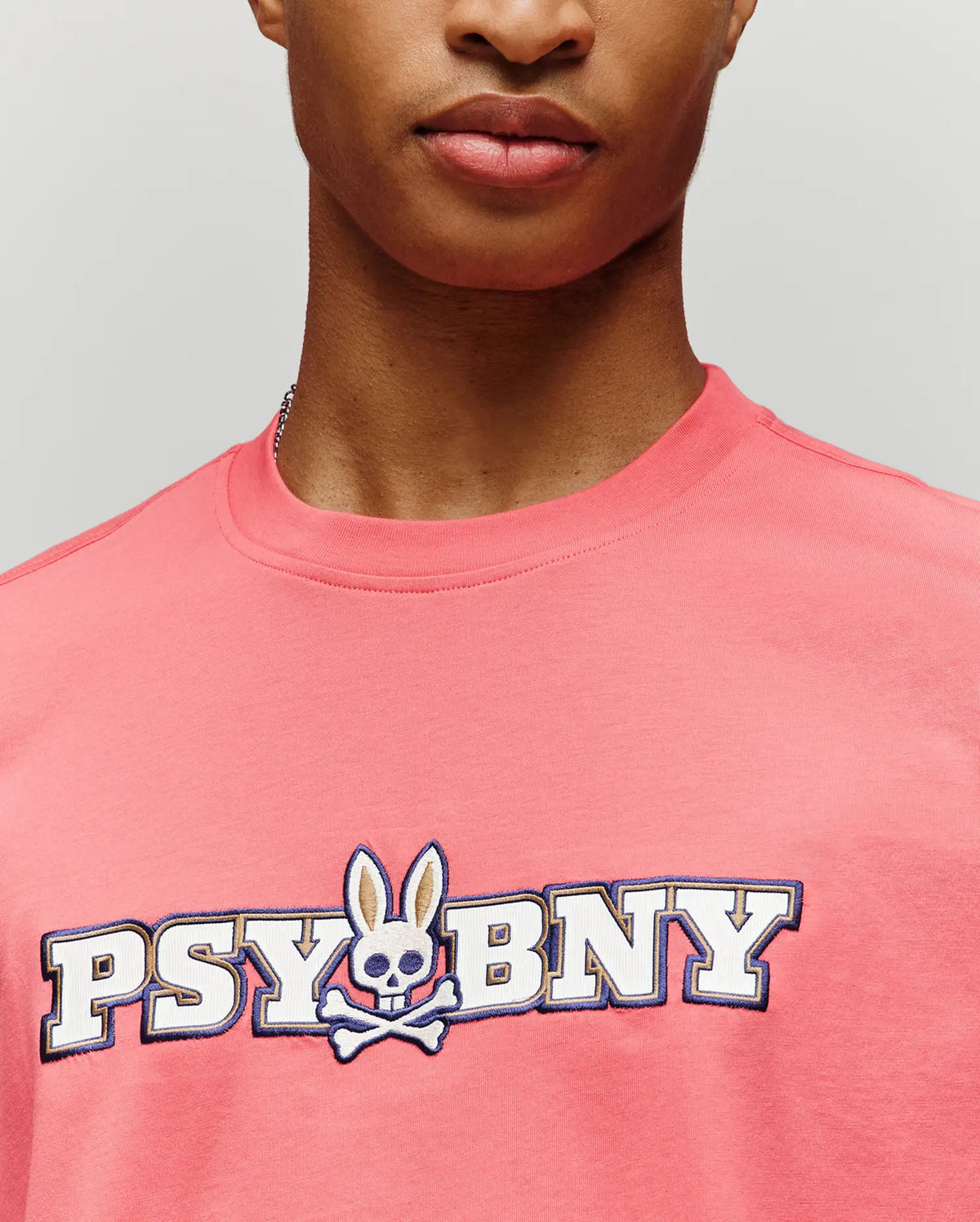 A person in the Psycho Bunny MENS WADE GRAPHIC TEE (MT0200105), a pink 100% cotton regular fit, featuring "PSYBNY" text and a bunny with skull face and crossbones, shown from shoulders to just below the nose.