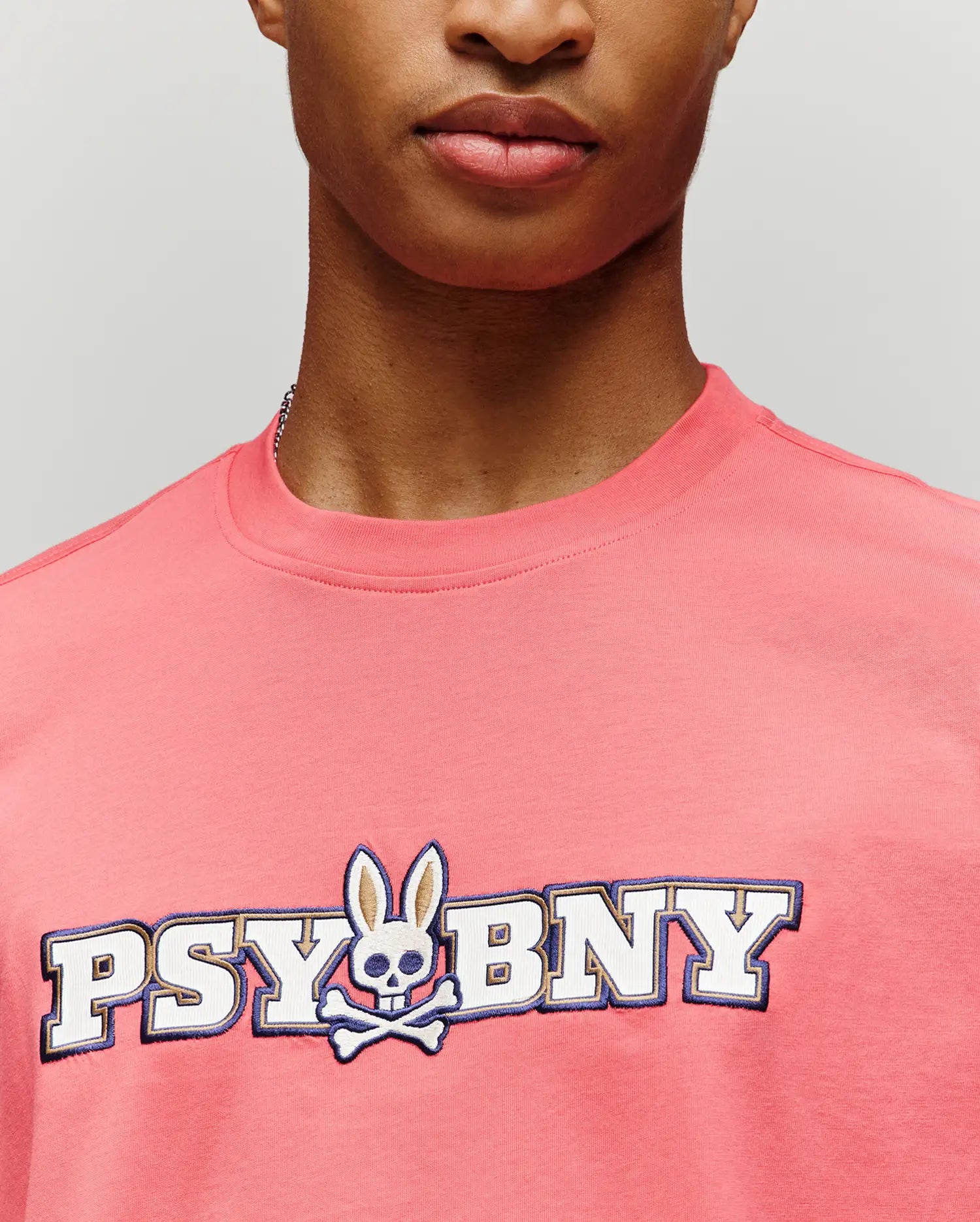 A person in the Psycho Bunny MENS WADE GRAPHIC TEE (MT0200105), a pink 100% cotton regular fit, featuring "PSYBNY" text and a bunny with skull face and crossbones, shown from shoulders to just below the nose.