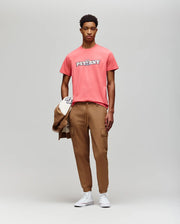 A young man faces the camera in a coral Psycho Bunny MENS WADE GRAPHIC TEE (MT0200105), tan cargo joggers, and white sneakers, holding a tan jacket and cap against a plain light gray background.