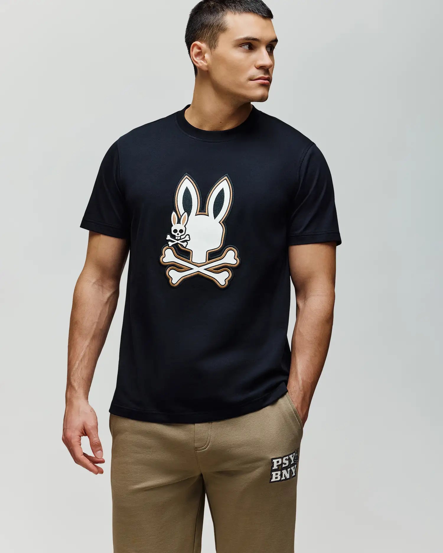 A man in a black Psycho Bunny MENS WADE GRAPHIC TEE (MT0200106) with an embroidered Bunny skull and crossbones, paired with tan 100% cotton shorts with white left leg text, stands against a light background, looking left.