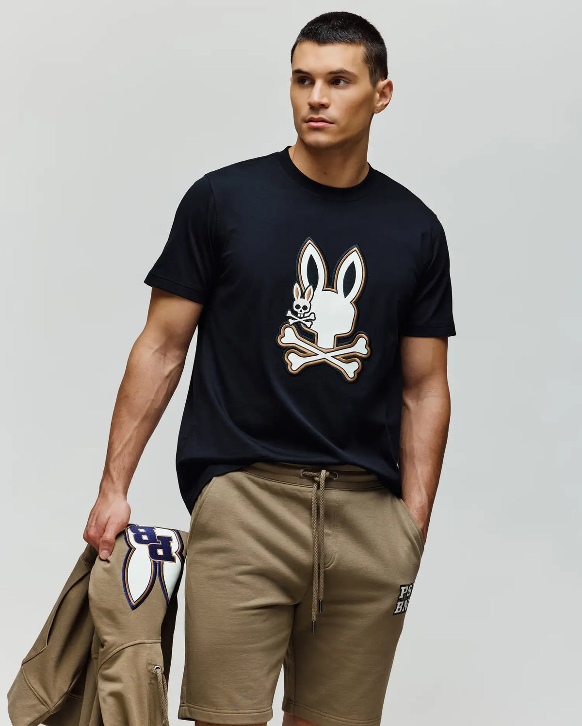 A man in a 100% cotton Psycho Bunny MENS WADE GRAPHIC TEE (MT0200106) with an embroidered Bunny and crossbones graphic, paired with tan shorts, stands against a plain light background holding a matching tan jacket.