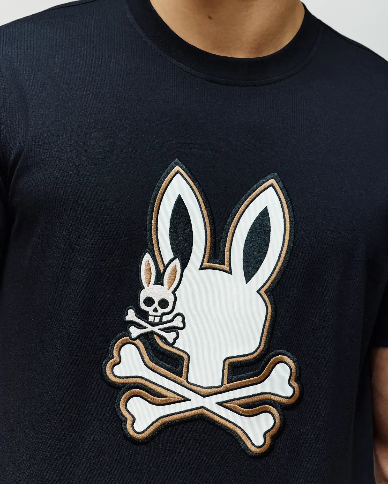 A person wears the Psycho Bunny MENS WADE GRAPHIC TEE - MT0200106, a black 100% cotton tee with a bold white bunny head and crossbones graphic plus an embroidered Bunny skull and crossbones on top.