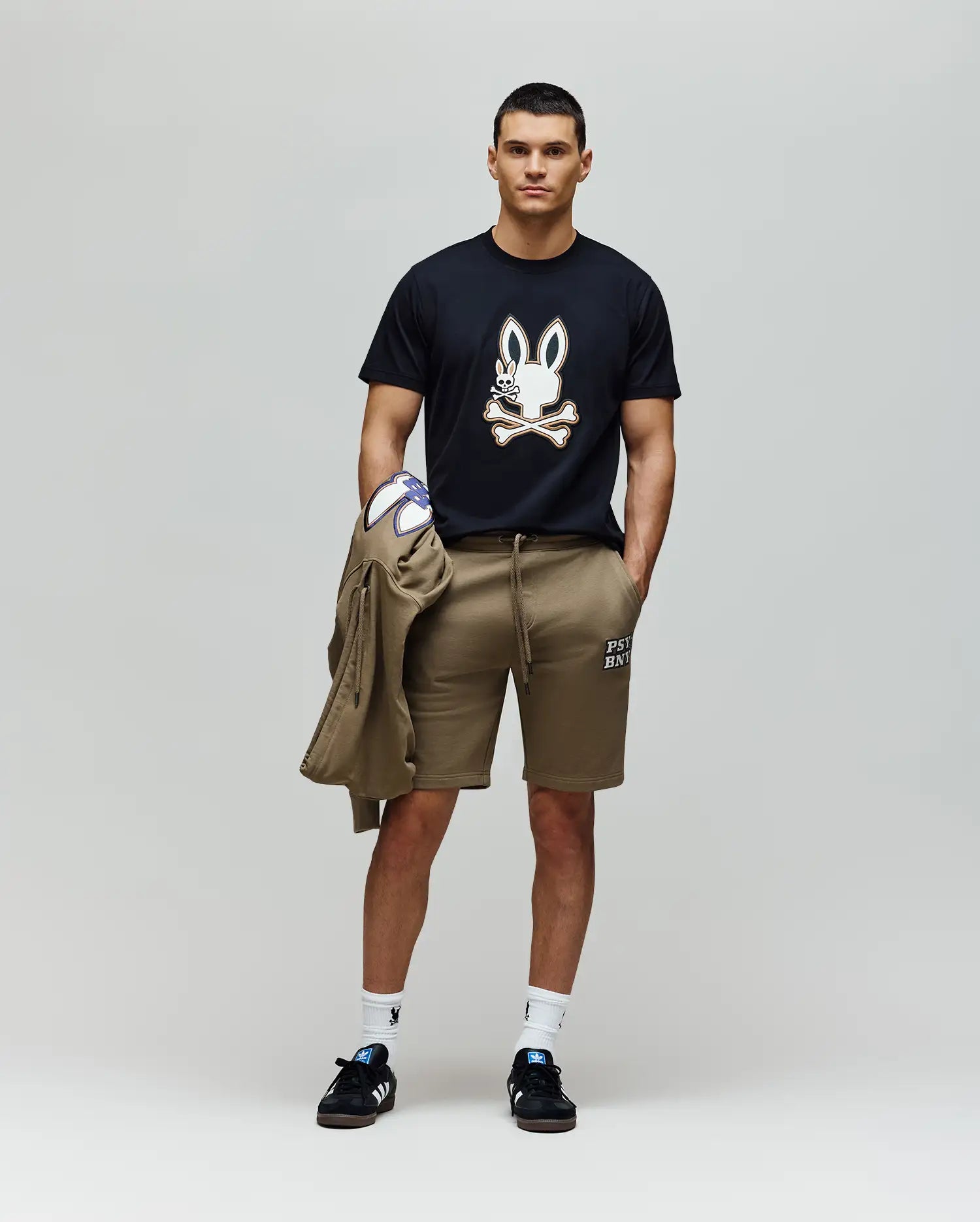 A person stands against a plain background wearing the Psycho Bunny MENS WADE GRAPHIC TEE - MT0200106 in black with an embroidered bunny on 100% cotton, paired with tan shorts, white socks, black sneakers, and holding a matching tan jacket.