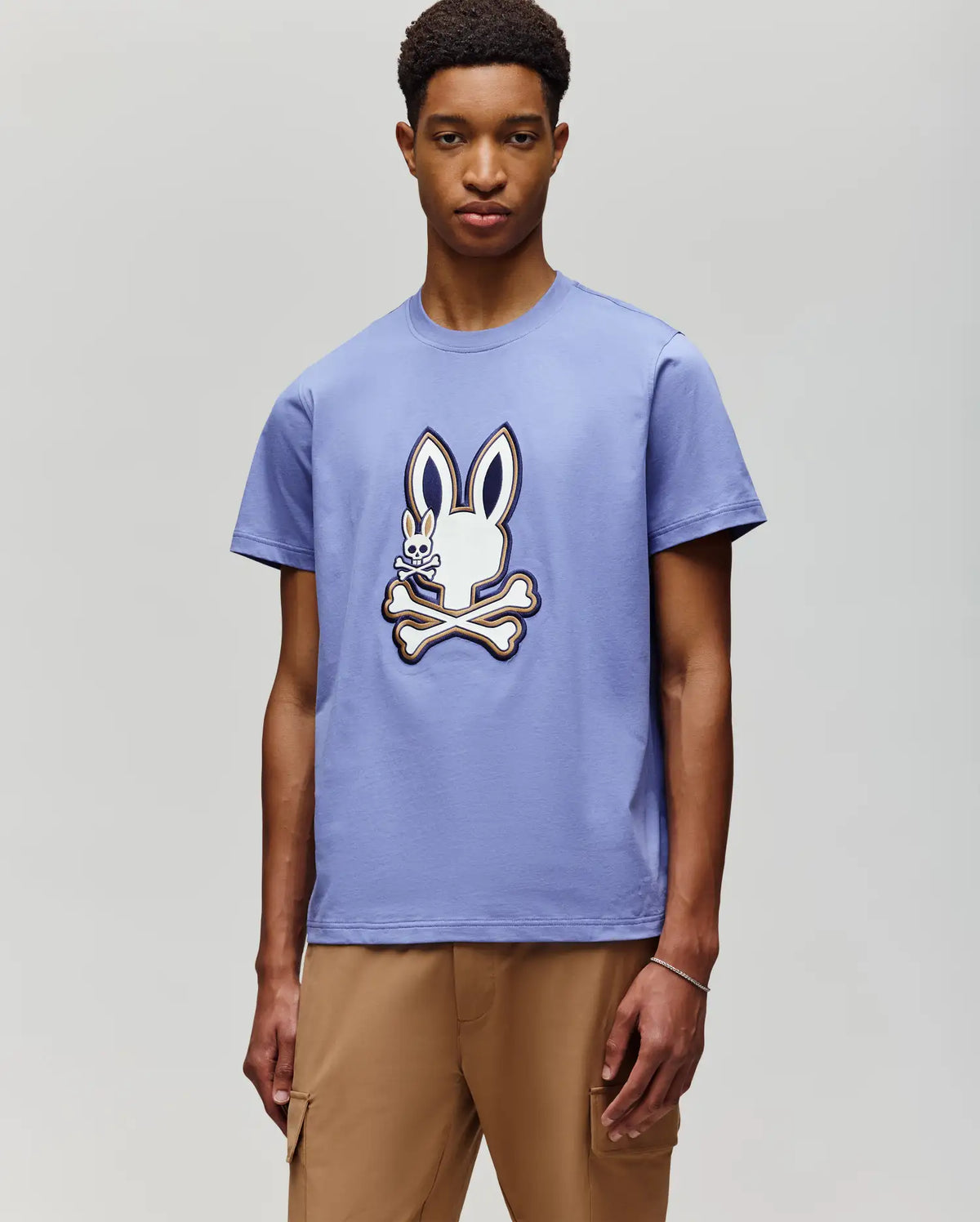 A young man with short hair models the Psycho Bunny MENS WADE GRAPHIC TEE (MT0200106), a 100% cotton shirt featuring an embroidered bunny above crossed bones, paired with tan pants against a light gray background.