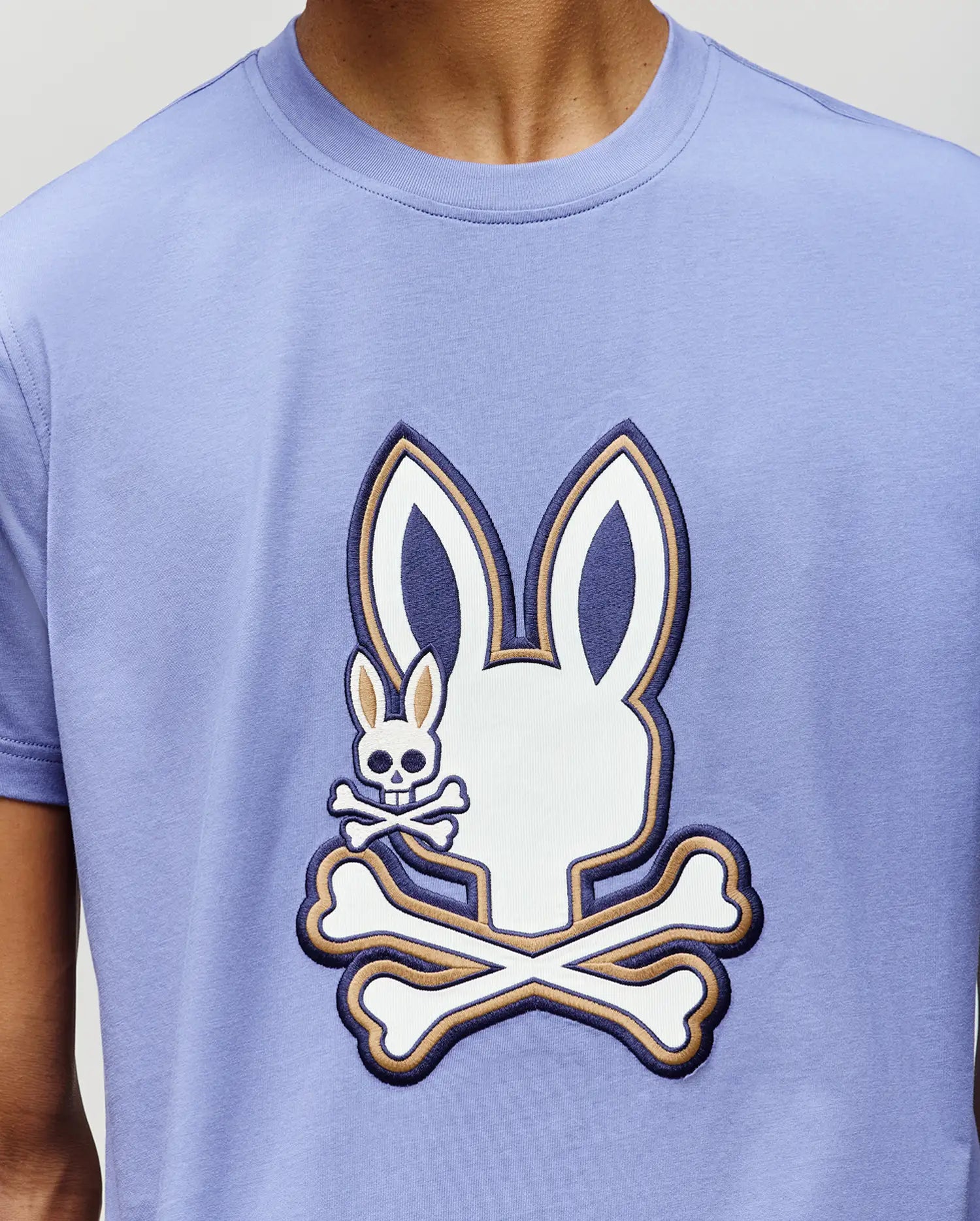 A person wearing the Psycho Bunny MENS WADE GRAPHIC TEE (MT0200106), a light purple 100% cotton shirt with a large white bunny skull and crossbones graphic, plus a smaller bunny skull and crossbones on the cheek.