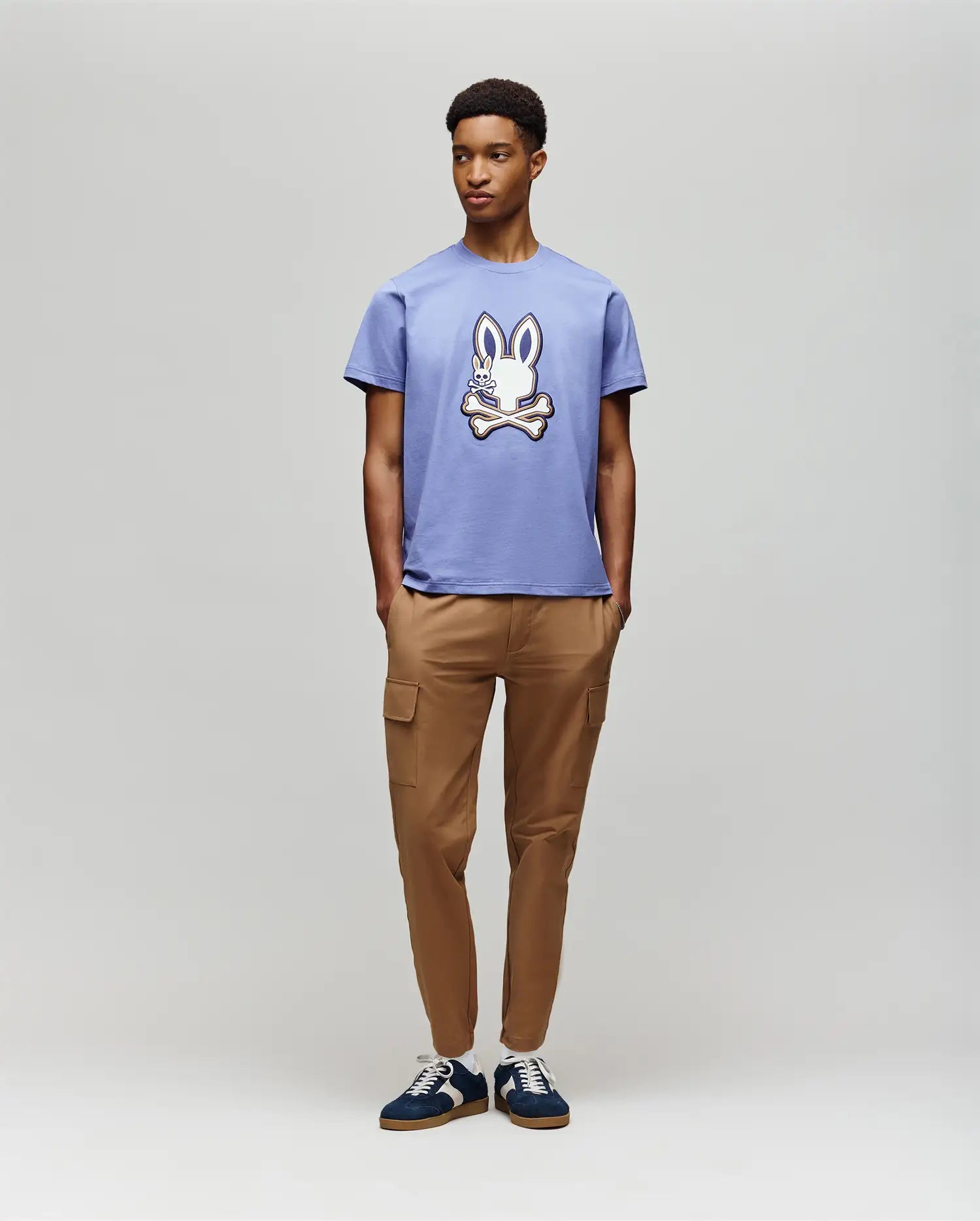A young man wears the Psycho Bunny MENS WADE GRAPHIC TEE - MT0200106 in lavender, featuring a 100% cotton build and an embroidered bunny, paired with tan cargo pants and dark sneakers against a plain background.