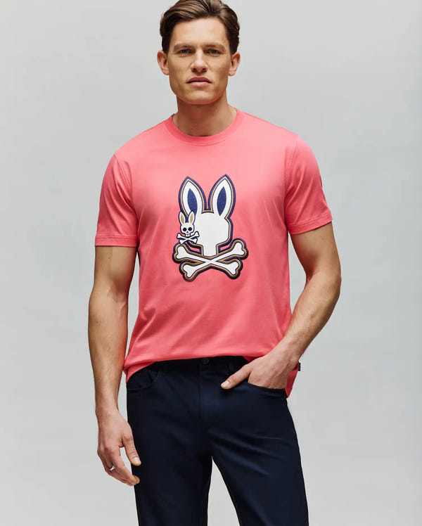 A man wears the Psycho Bunny MENS WADE GRAPHIC TEE - MT0200106, a pink 100% cotton shirt with a cartoon bunny and crossbones graphic on the front, paired with dark pants against a plain light gray background.