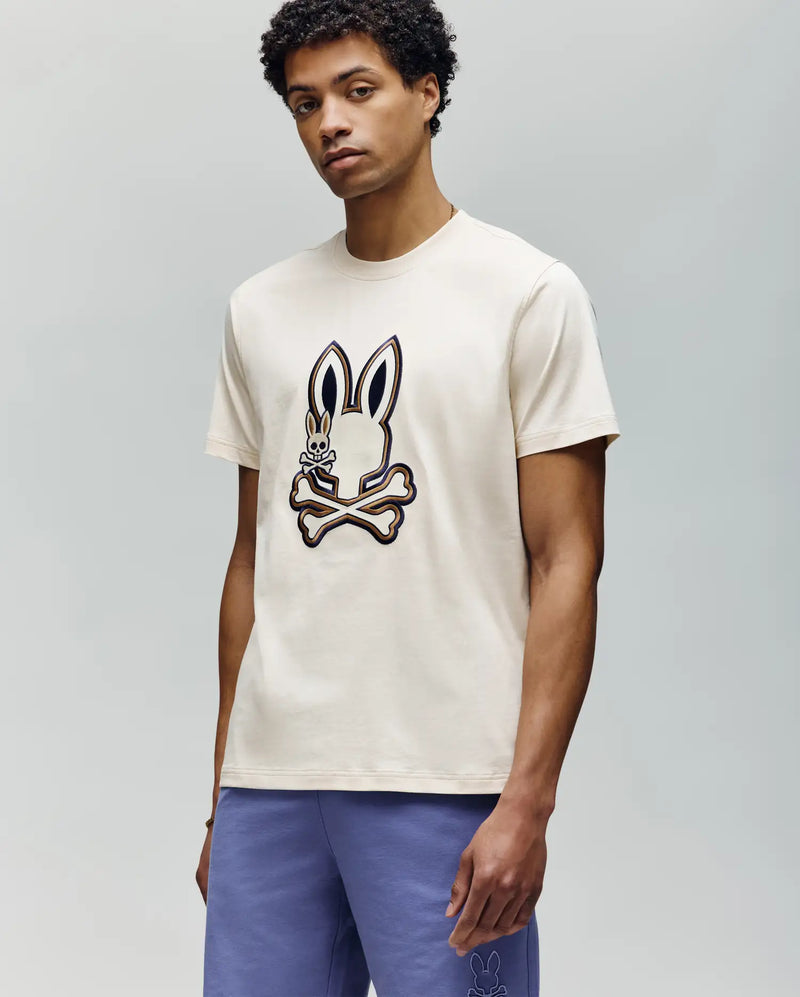 A man wearing the Psycho Bunny MENS WADE GRAPHIC TEE - MT0200106, a cream tee with a large embroidered bunny and crossbones on the front, paired with purple shorts, stands against a plain light gray background.