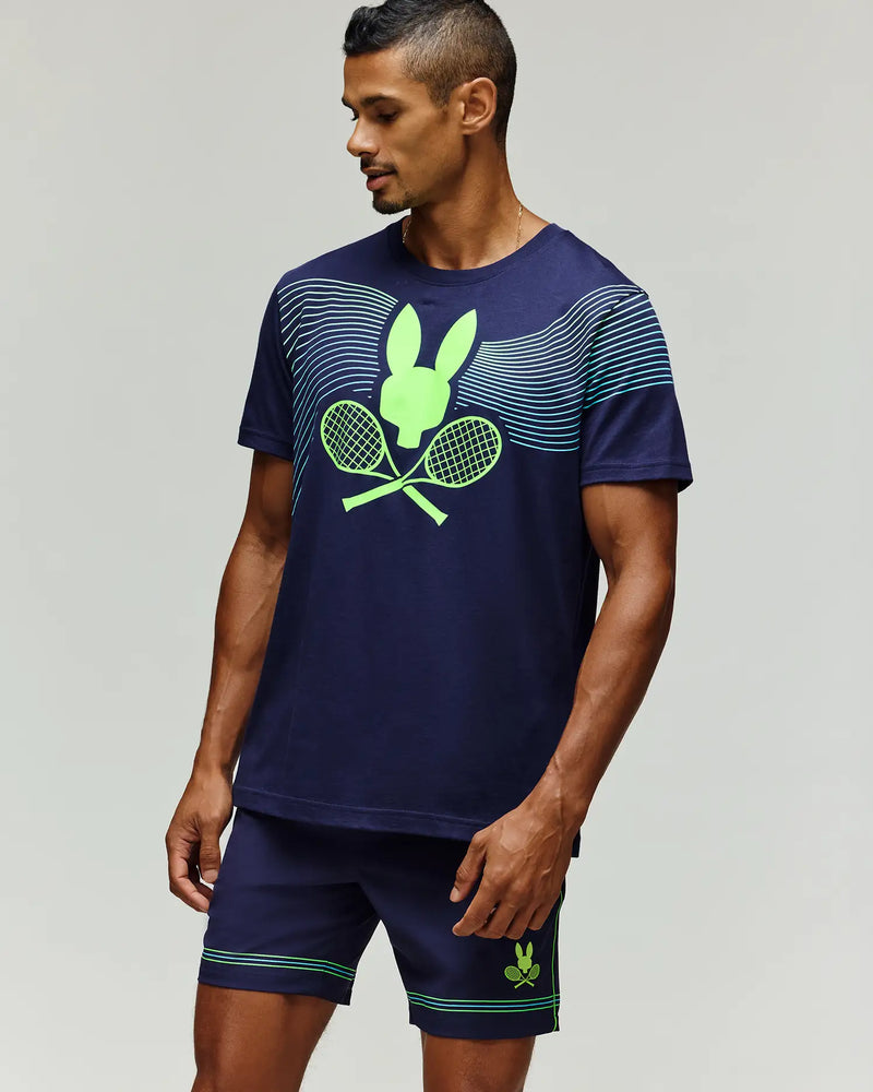 A man wears a Psycho Bunny TENNIS BALL CREW MENS TEE ENGLISH (MT0200113), a navy blue Pima cotton tee with neon green crossed rackets and bunny logo, paired with shorts, standing against a plain light background.