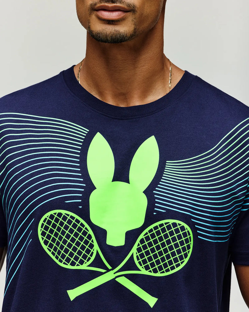 A man wears the Psycho Bunny Tennis Ball Crew Men's Tee (MT0200113) in navy blue, made from soft Pima cotton, with a neon green bunny head above crossed tennis rackets and light blue radiating lines; only his lower face and upper torso show.