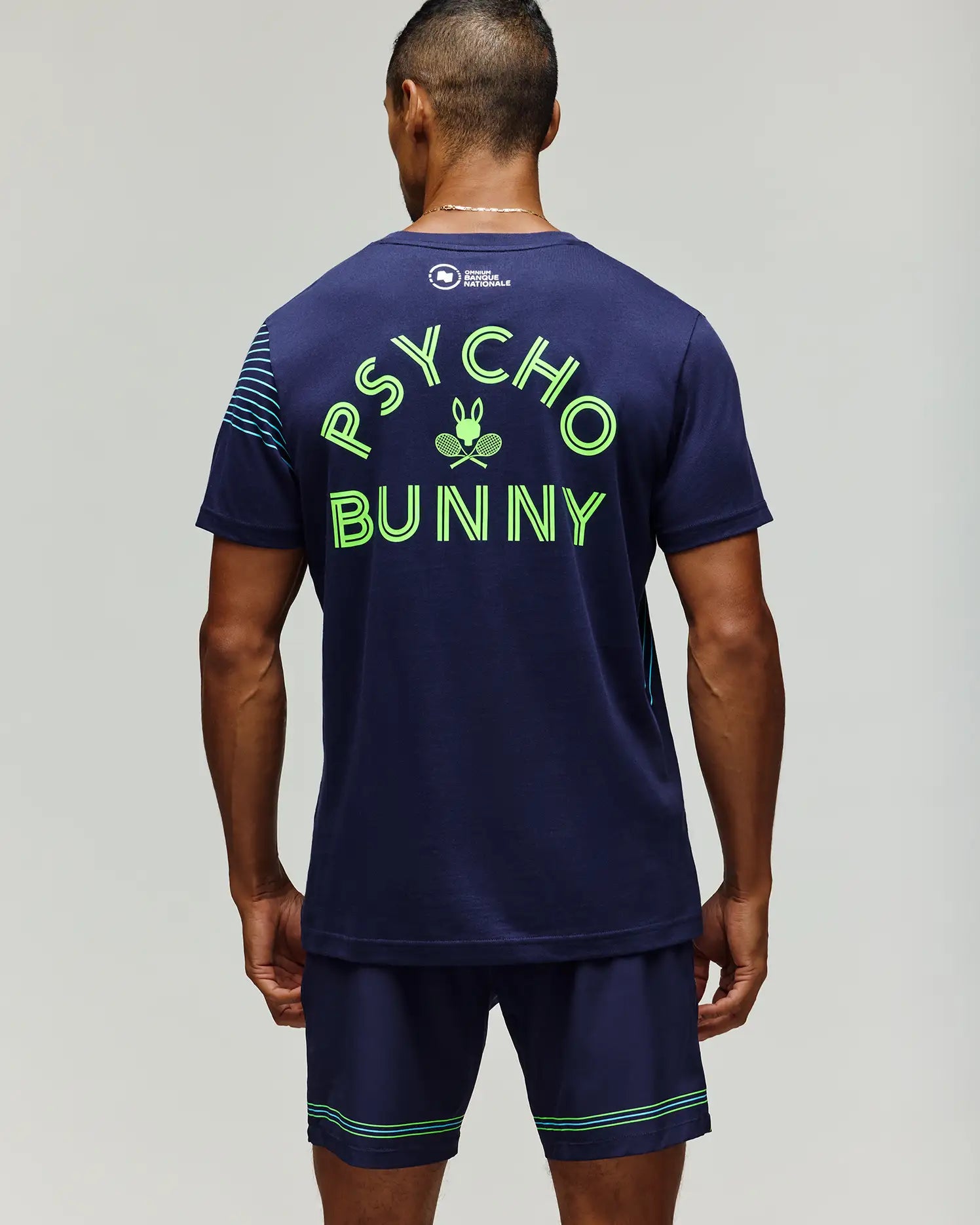 A man faces away in Psycho Bunny's TENNIS BALL CREW MENS TEE FRENCH (MT0200114), a navy Pima cotton tee featuring neon green "PSYCHO BUNNY" text and bunny logo, paired with coordinating navy shorts with green stripes.