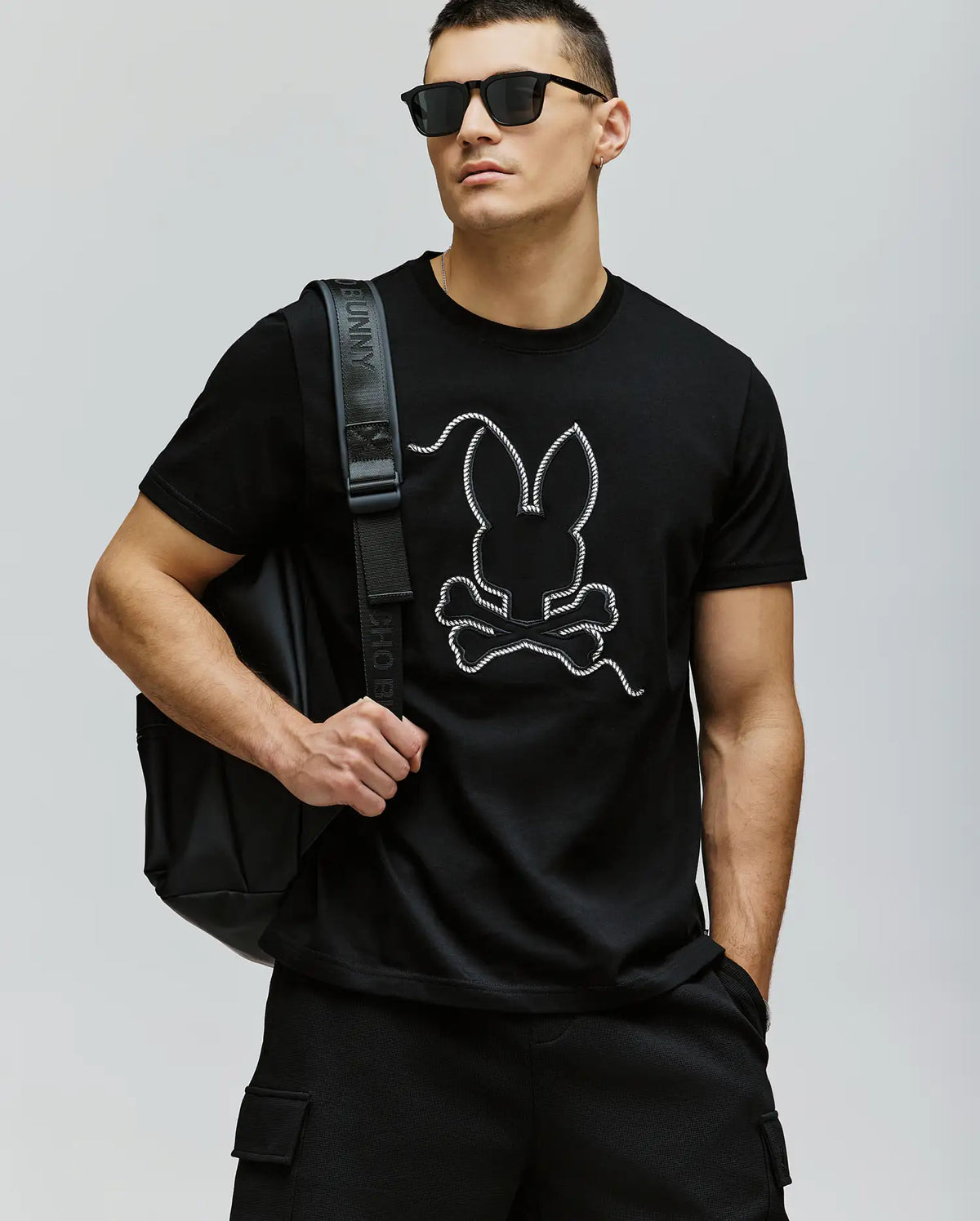 A man wearing sunglasses, black shorts, and a backpack poses confidently with one hand in his pocket, sporting the Psycho Bunny MENS CYRUS GRAPHIC TEE (MT0200115), a black 100% Pima cotton tee with a stylized bunny and crossbones.