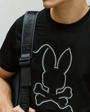 A person wears the Psycho Bunny MENS CYRUS GRAPHIC TEE (MT0200115), a 100% Pima cotton shirt featuring a white bunny and crossbones outline, paired with a black shoulder strap bag. The cropped image highlights a sailor-inspired vibe.