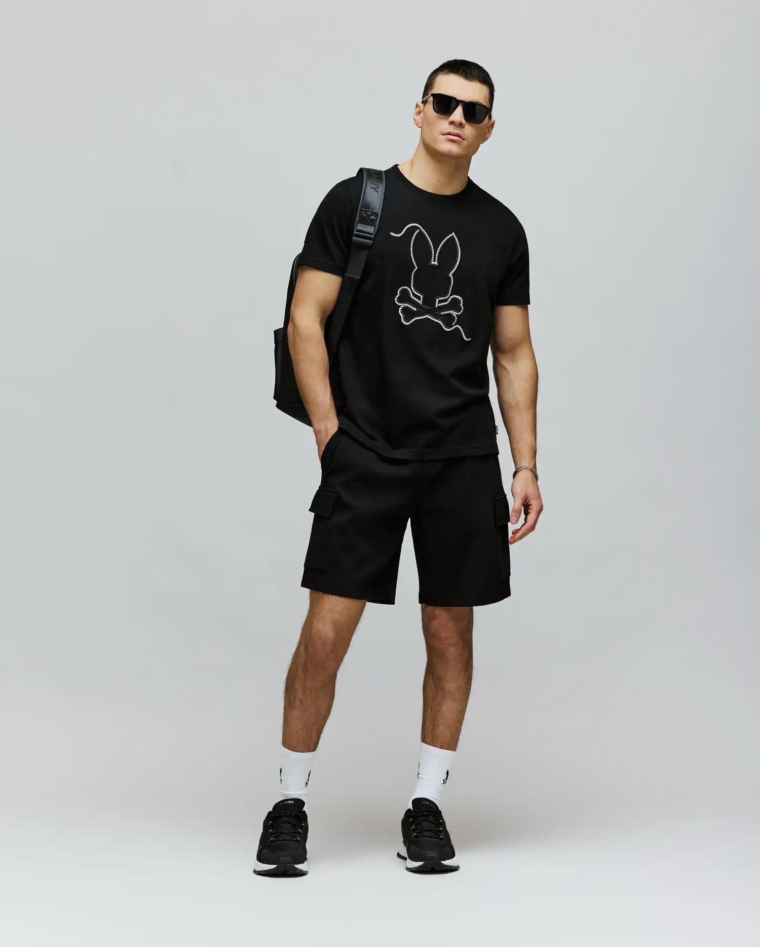 A man in sunglasses, a Psycho Bunny MENS CYRUS GRAPHIC TEE (MT0200115) made of 100% Pima cotton, black shorts, black sneakers, white socks, and a black backpack stands against a plain light background.