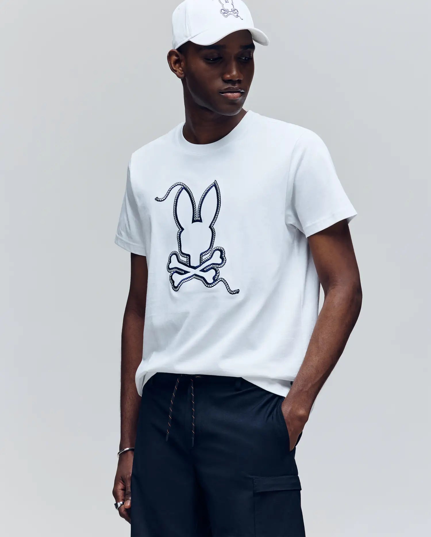 A man wears the Psycho Bunny MENS CYRUS GRAPHIC TEE (MT0200115), a soft Pima cotton shirt with a large outlined bunny head above crossed bones, paired with dark pants as he looks left against a plain background.
