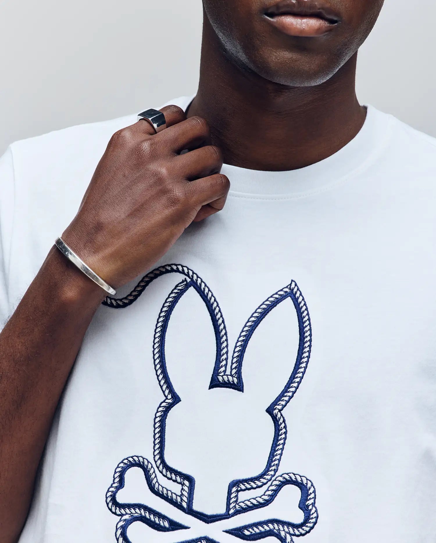 Wearing the Psycho Bunny MENS CYRUS GRAPHIC TEE (MT0200115), featuring a rope-outline bunny and crossbones, a person holds their collar. The image highlights their silver bracelet, black ring, and shows only the lower face and upper torso.