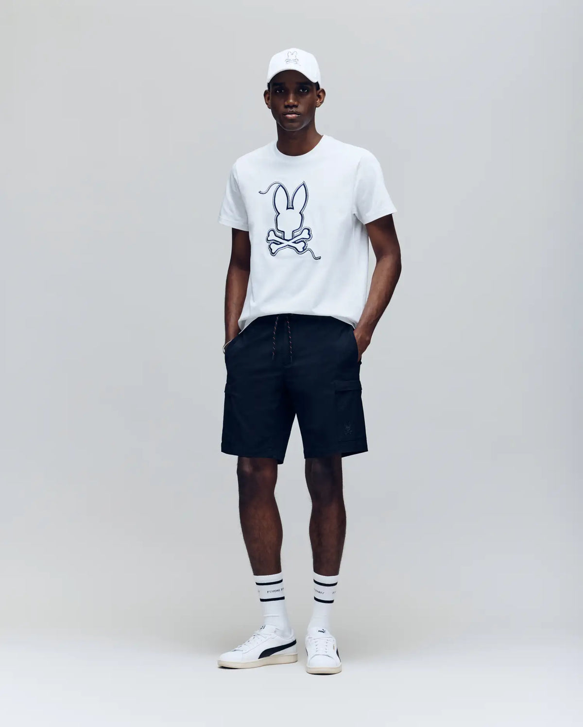 A man stands against a plain background wearing the Psycho Bunny MENS CYRUS GRAPHIC TEE - MT0200115, black shorts, a white cap, striped white socks, and white sneakers with his hands in his pockets.