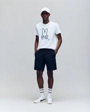 A man stands against a plain background wearing the Psycho Bunny MENS CYRUS GRAPHIC TEE - MT0200115, black shorts, a white cap, striped white socks, and white sneakers with his hands in his pockets.