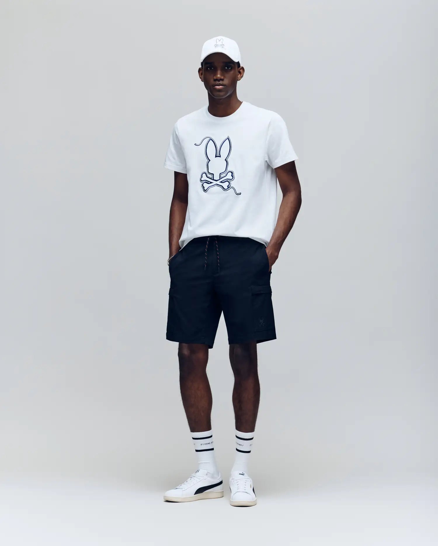 A man stands against a plain background wearing the Psycho Bunny MENS CYRUS GRAPHIC TEE - MT0200115, black shorts, a white cap, striped white socks, and white sneakers with his hands in his pockets.