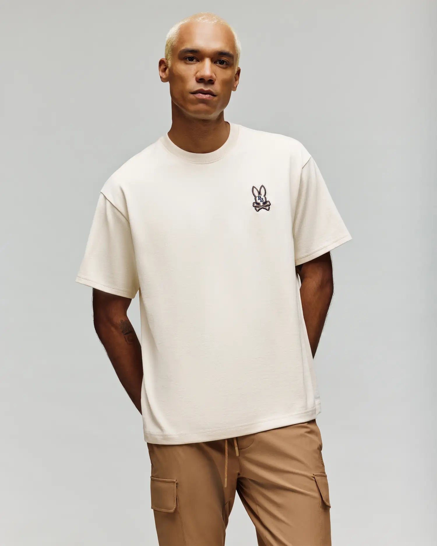 A person with short blond hair wears the Psycho Bunny MENS WADE OVERSIZED FIT CHEST TEE (MT0200152) in off-white, featuring a small cartoon bunny emblem, paired with tan cargo pants against a plain light background.