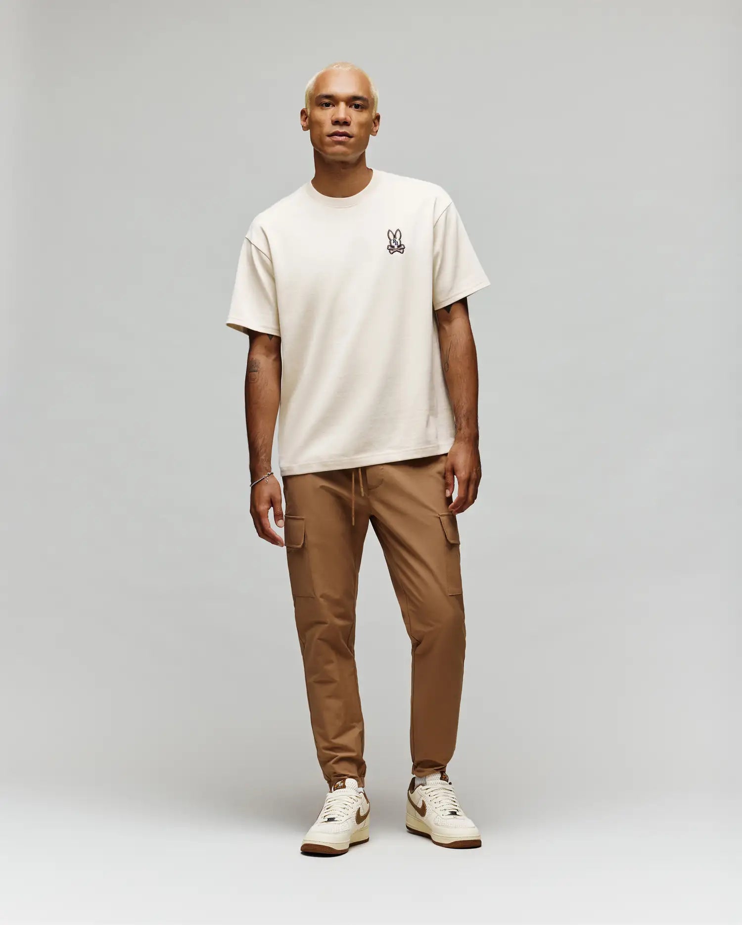 A person with light blond hair wears a Psycho Bunny MENS WADE OVERSIZED FIT CHEST TEE (MT0200152) in beige, styled with tan cargo pants and beige sneakers, standing against a plain light gray background.