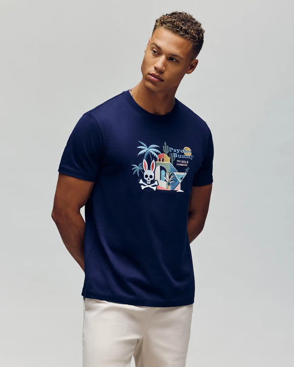 A man stands with his hands behind his back, looking to the side, wearing light-colored pants and a Psycho Bunny MENS SHANE GRAPHIC TEE (MT0200175) featuring a navy blue color and a vibrant embroidered cartoon rabbit with tropical accents.