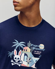 A person wears the Psycho Bunny MENS SHANE GRAPHIC TEE (MT0200175), a navy summer tee in soft Pima cotton with colorful embroidered tropical graphics—palm tree, cactus, building, cocktail, and bunny skull logo. Text: "Psycho Bunny THE BOLD STANDARD.